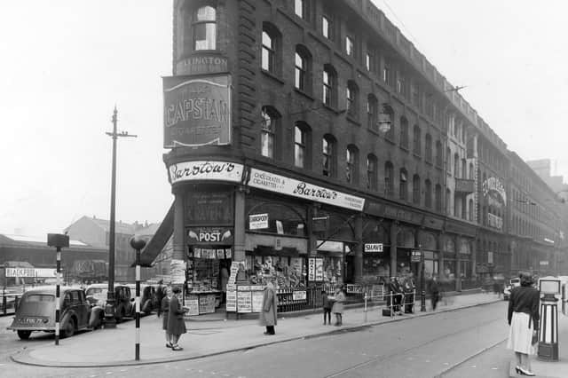 The junction of Aire Street and Wellington Street on City Square in June 1939. The focus of the photo is the premises of W Barstow, tobacconists, newsagents, confectioners, hairdressers and L Barstow, chiropody. At the newsagent end of the shop is an extensive display of magazines and newspaper billboards with clear contemporary headlines. There is a neon sign for Capstan cigarettes and below this above the shop doorway an advert for Craven A cigarettes, saying 'for your throat's sake smoke Craven A.' The edge of the railway station can be seen down Aire Street and there is a long line of parked cars along Wellington Street.