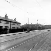  Belle Isle Road looking south towards an iron railway bridge in November 1950. On the right of the road there is a brick industrial building with a wall and iron fence running along the pavement. There is a woman walking along the pavement and a car parked at the kerb. On the left are two rows of stone terraced houses. In the road are tram lines and a traffic island.At the left hand kerb stands a window cleaners barrow.