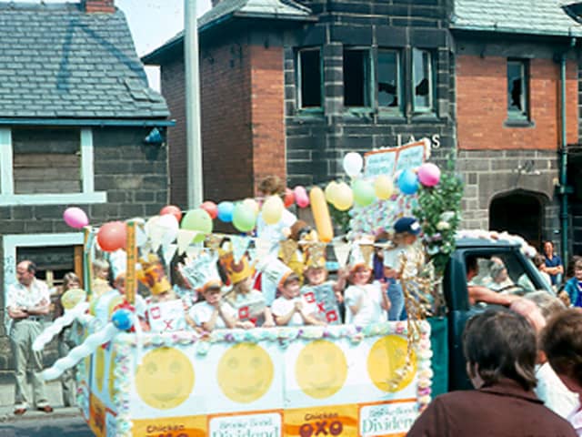 A decorated float parading along Upper Town Street during the Bramley Carnival of 1976, with people lining the streets to watch. The carnival had been a regular event in the early part of the 20th century but the 1976 one was the first for many years. Buildings in the background numbered 216 - 218 are boarded up or have windows broken and are scheduled for demolition.