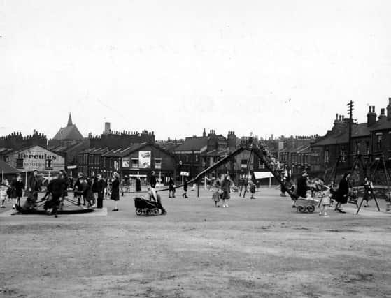 View across the playground of Beckett Street recreation ground in June 1949. Adults with children, some in prams, play on the swings, slide and roundabout. Behind are terraced houses. One gable end is painted with an advertisement for 'Hercules Modern Cycles'.