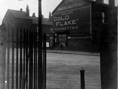 Hunslet Moor railway crossing on Beza Street. Metal railings, a streetlamp and a bollard are in the forgeround. Number 48 Parkside sweet stores is opposite with a large advertisement for Wills Gold Flake cigarettes above. Prospect Street and Prospect Terrace are partially visible. Pictured in May 1929.