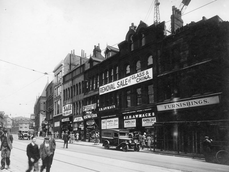 New Briggate in July 1931. Number 8 to 10 New Briggate is J.H. Awmack Ltd Glass and China dealers with works vehicle parked outside. Next to this at number 12 is Thomas Whiteley and Daughters hairdressers with chiropodists above. Number 14 is Fit-rite shoe company, then at 16 Mentel Ltd furriers. Pitman College can then be seen with basement shop to let below. Beyond Harrison Street can be seen the Assembly Rooms which was built in 1898 as a concert hall.