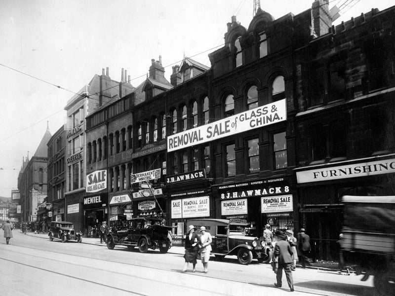New Briggate in July 1931. Numbers 8 to 10 J.H. Awmack Ltd, glass and china dealers with works vehicle and other vehicles parked outside. Number 12 is Thomas Whiteley and Daughters hairdressers with chiropodist above. At 14 fit-rite shoe company. Number 16 is Mentel Ltd, furriers. Number 20 is Pitmans College with Basement shop to let below.