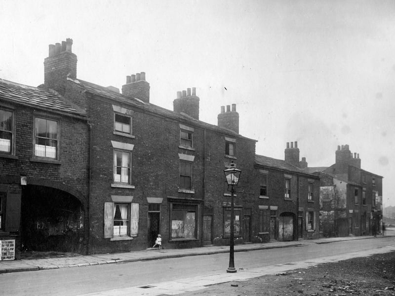 Bridge Street prior to demolition. Brick terraced houses with sash windows, shutters & net curtains. Archways to courtyard. One building has cinema adverts underneath window. A small girl stands in doorway of another building. Lamp post in foreground. Pictured in July 1931.