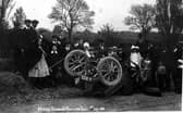 A postcard with 'Motor Car smash at Rawdon' on it. Group of people in the background, wearing clothes typical of the period.  Pictured in June 1909.