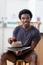 Josh Tedeku as Cory Maxson, holding a script and a fedora.