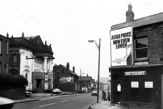 A tattooist on the corner of Domestic Street offers 'expert tattoo removal' and 'painless ear piercing'. An advertising hoarding for Asda supermarket covers the wall. On the left of the picture is Holbeck Mills Cash and Carry Carpet Warehouse, formerly the United Methodist Chapel. The junctions with Ingram View and Willoughby Terrace are at either side.