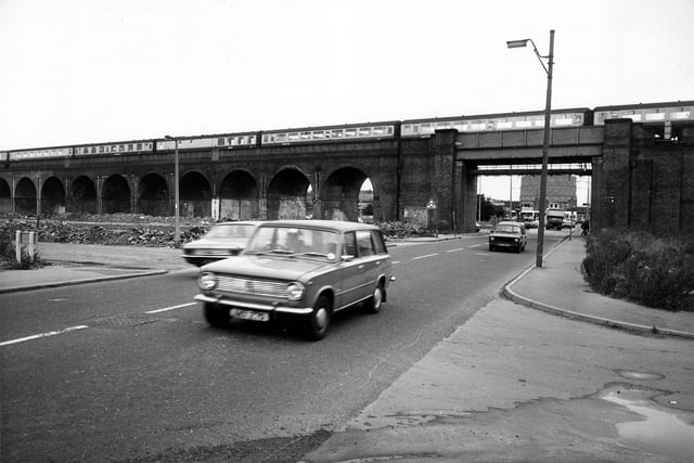 Tthe railway viaduct crossing Domestic Street in February 1980. An Inter-City train with many carriages is passing overhead. Cars can be seen on the road. A gasometer is visible in the distance through the archway.