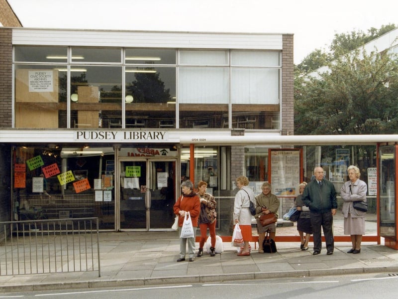 Pudsey Branch Library on Church Lane which was built in 1963 and opened on the in January 1964. Various notices are displayed in the windows. A queue of people are waiting at a bus shelter in front.