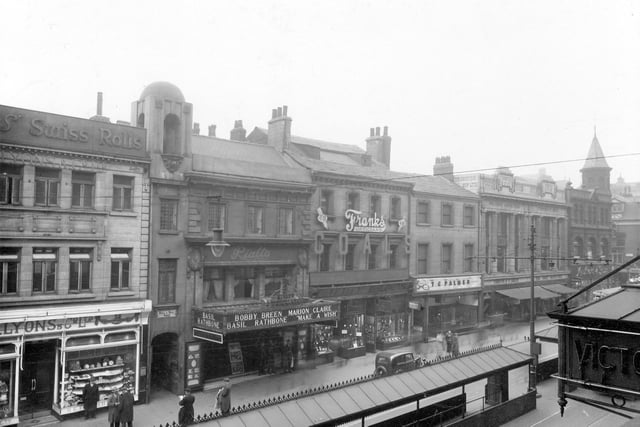 Shops on Briggate in March 1938. Pictured, from left, is Lyons and Co. Ltd; Bull and Bell yard; Rialto Cinema; Frank's Optician's; T.C.Palmer; Thornton's Ltd; and Mansfield and sons Ltd. Down the middle of the road can be seen bus/tram shelters.