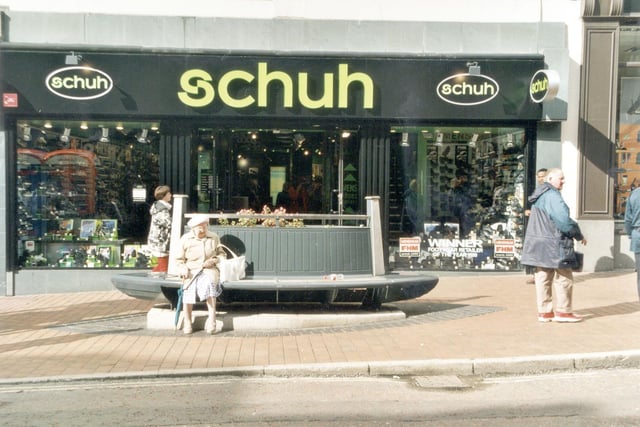 A lady sits on a bench in front of 'Schuh' shoe shop on Briggate.