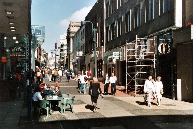 A busy Albion Street in September 1999. People sit on the chairs outside the sandwich shop. Leeds Shopping Plaza is to the left and in particular, JJB Sports Superstore. On the right is The Link, mobile phone shop, Waterstones, booksellers and Specsavers, opticians.