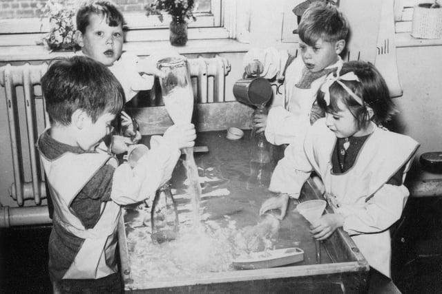 Four children at Bentley Lane Infants school engaged in water play activities. They are pouring water from jugs into bottles and funnels, and then emptying them.