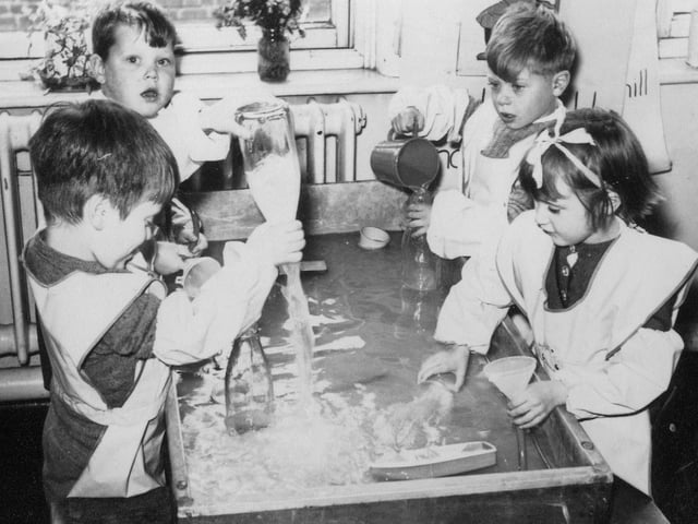 Four children at Bentley Lane Infants school engaged in water play activities. They are pouring water from jugs into bottles and funnels, and then emptying them.
