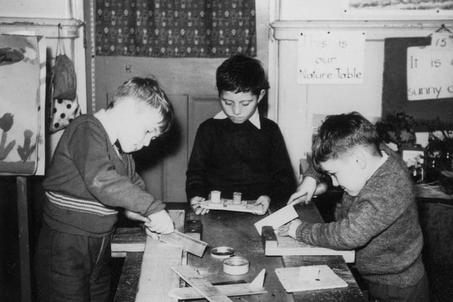 Three boys are absorbed in woodworking activities. Two of them are working with tenon saws, whilst the third is holding a previously made model boat. At the front is a a wooden model of an aeroplane, and at the back, right, can be seen part of the nature table.