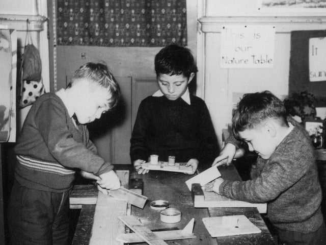 Three boys are absorbed in woodworking activities. Two of them are working with tenon saws, whilst the third is holding a previously made model boat. At the front is a a wooden model of an aeroplane, and at the back, right, can be seen part of the nature table.