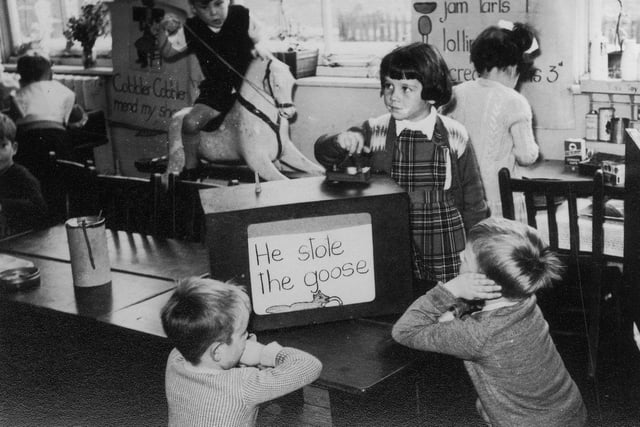Children engaged in various activities. At the front, three children are gathered around a mock-up of a television set, whilst behind them a boy plays on a rocking horse, and a girl plays in a 'shop' area.