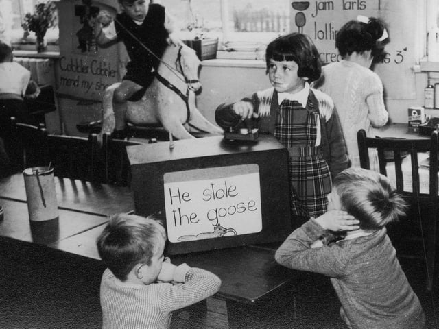 Children engaged in various activities. At the front, three children are gathered around a mock-up of a television set, whilst behind them a boy plays on a rocking horse, and a girl plays in a 'shop' area.