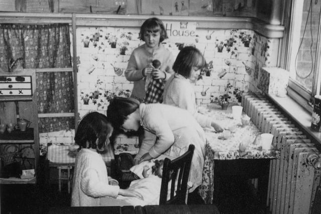 Four girls are pictured playing in the 'home corner'.  Two are playing with a doll in a crib, and two are standing by a table set with small cups and saucers.