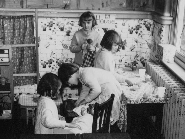 Four girls are pictured playing in the 'home corner'. Two are playing with a doll in a crib, and two are standing by a table set with small cups and saucers.