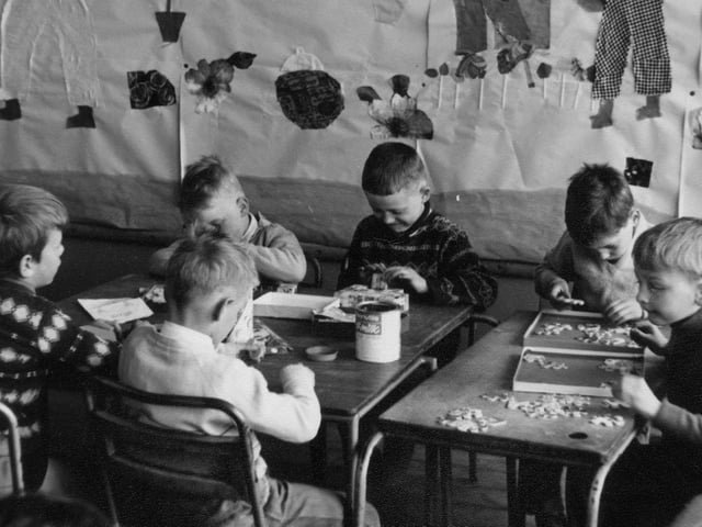 A group of six boys, all absorbed in completing jigsaw puzzles. Behind them is a wall collage featuring flowers and figures.