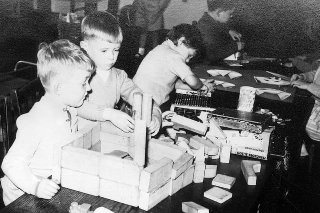 A group of children engaged in various practical activities. Two boys at the front are building with wooden blocks, in the centre is a child using a counting frame, and at the back the activity would appear to be origami.
