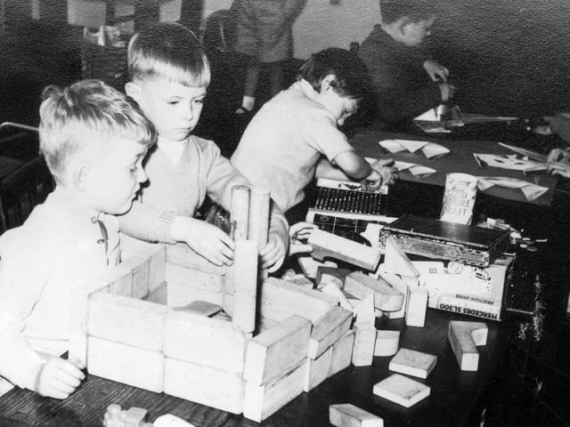 A group of children engaged in various practical activities. Two boys at the front are building with wooden blocks, in the centre is a child using a counting frame, and at the back the activity would appear to be origami.