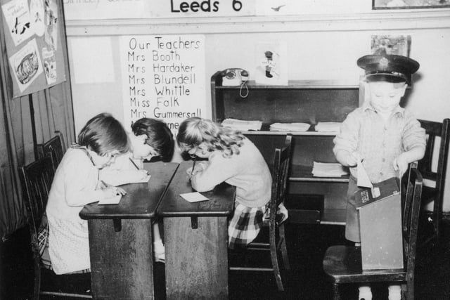 Four children involved in dressing up and role play. The child in the right is wearing a postman's hat and posting a letter into a letter box. The three children on the left are writing letters and addressing envelopes, presumably to be delivered by the junior postman on the right.