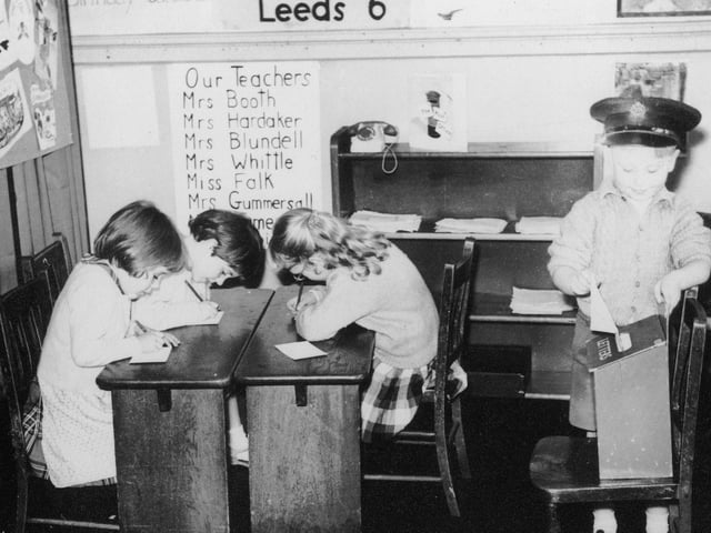 Four children involved in dressing up and role play. The child in the right is wearing a postman's hat and posting a letter into a letter box. The three children on the left are writing letters and addressing envelopes, presumably to be delivered by the junior postman on the right.