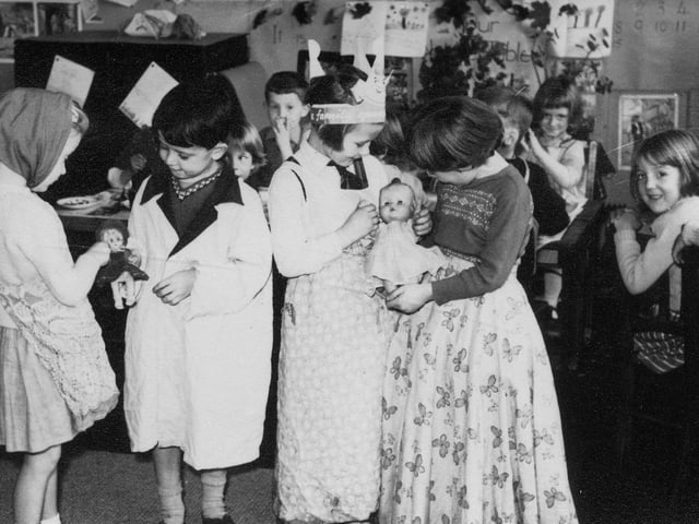 Four children who have been dressing up, and are playing with dolls. Several children are sitting behind them, looking on.