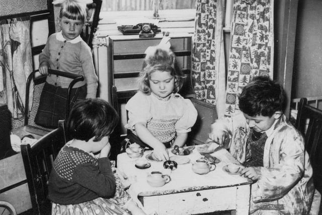 Three children are pictured holding a pretend tea-party. They are using miniature cups and models of food. To the left, a girl pushing a cart or pram looks on.
