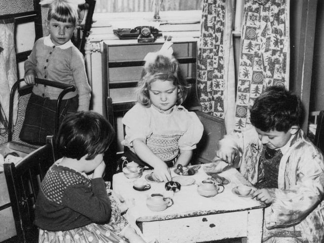 Three children are pictured holding a pretend tea-party. They are using miniature cups and models of food. To the left, a girl pushing a cart or pram looks on.