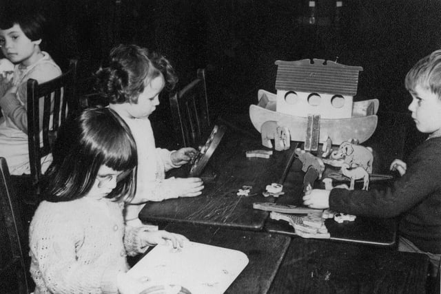 A group of children playing at wooden tables. On the right a boy is playing with a model of Noah's Ark, complete with wooden animals. The girl opposite him is playing with what may be a wooden toy clock.