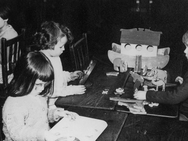 A group of children playing at wooden tables. On the right a boy is playing with a model of Noah's Ark, complete with wooden animals. The girl opposite him is playing with what may be a wooden toy clock.