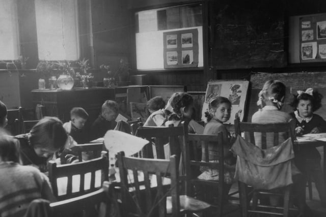 Children working in a classroom. Most appear to be absorbed in drawing or writing, and there are pictures either side of the blackboard, which shows a sketch of part of the world map. On the left are plants on a cabinet, possibly part of a 'nature corner'.