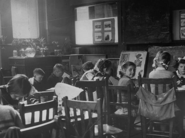 Children working in a classroom. Most appear to be absorbed in drawing or writing, and there are pictures either side of the blackboard, which shows a sketch of part of the world map. On the left are plants on a cabinet, possibly part of a 'nature corner'.