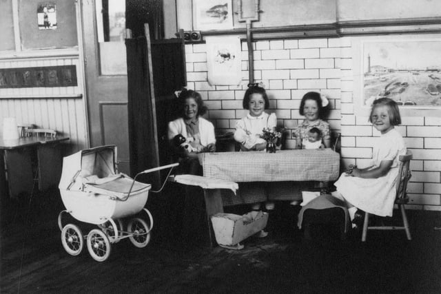Pictured are four girls with dolls, posing for the camera. By the table at which they are seated are a miniature crib, bed, and ironing board, and to the left is a pram, and some small wooden tables.