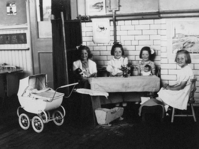 Pictured are four girls with dolls, posing for the camera. By the table at which they are seated are a miniature crib, bed, and ironing board, and to the left is a pram, and some small wooden tables.