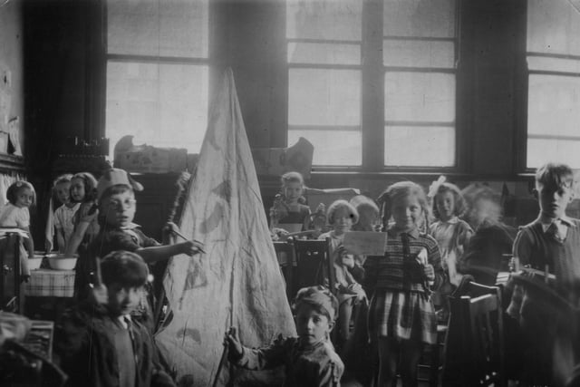 A group of children in a classroom. A variety of activities are taking place, including dressing up, painting, modelling, and writing. A girl at the front is holding up a written note, partially legible, which indicates the children are in 'Class 2'. A model 'wigwam' is on the left.
