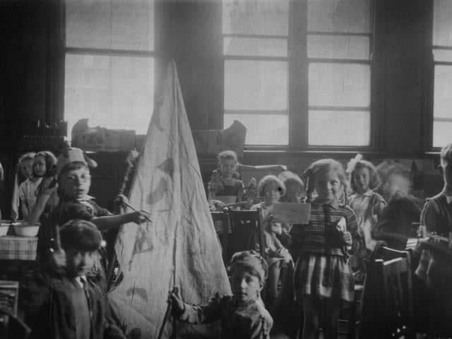 A group of children in a classroom. A variety of activities are taking place, including dressing up, painting, modelling, and writing. A girl at the front is holding up a written note, partially legible, which indicates the children are in 'Class 2'. A model 'wigwam' is on the left.
