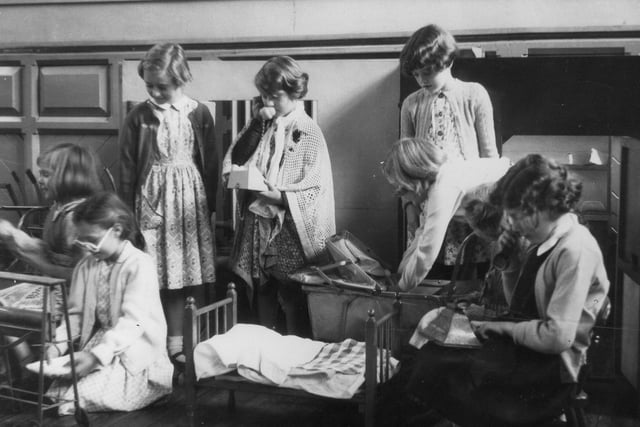 A group of girls engaged in dressing-up and role-play activities. One can be seen, right placing a doll in a pram, whilst two others are pretending to use telephones. A miniature bed is in the centre of the photograph.
