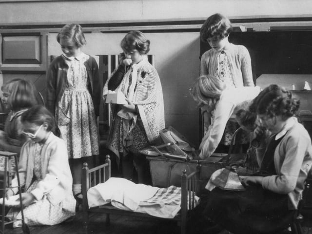 A group of girls engaged in dressing-up and role-play activities. One can be seen, right placing a doll in a pram, whilst two others are pretending to use telephones. A miniature bed is in the centre of the photograph.