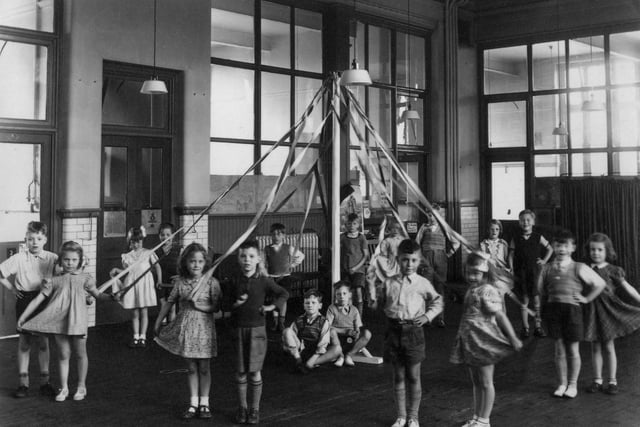 A group of children who have been practising a maypole dance, associated with May Day celebrations, and often performed by local school children. The dance involved weaving of the ribbons to form patterns around the pol. The children here are dancing in pairs, and two boys can be seen sitting sat the base of the pole to support it.