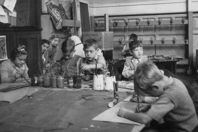 A group of children are pictured working on a painting, while two can be seen looking round the corner. Brushes can be seen in jars and jugs on the table, plus pencils in a pot. Behind the main group, a boy is playing with some puzzles. Pegs for coats and bags are by the rear wall.