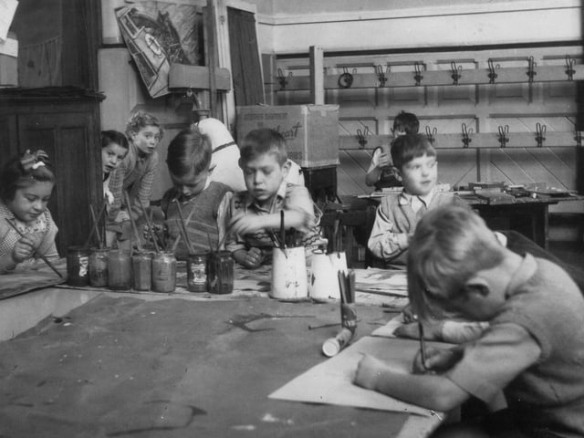 A group of children are pictured working on a painting, while two can be seen looking round the corner. Brushes can be seen in jars and jugs on the table, plus pencils in a pot. Behind the main group, a boy is playing with some puzzles. Pegs for coats and bags are by the rear wall.