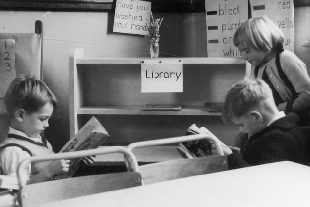 Two boys and a girl in the 'library corner'. The shelves are largely empty of books. and there are posters pinned to the wall behind, one naming a variety of colours.