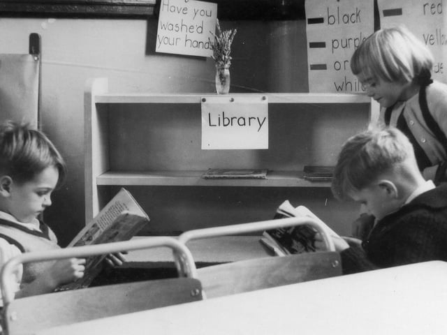 Two boys and a girl in the 'library corner'. The shelves are largely empty of books. and there are posters pinned to the wall behind, one naming a variety of colours.