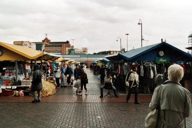 A view of the outdoor market is taken from just outside the doors of the indoor market and shows a row of stalls leading down to the City Bus Station. In the distance can be seen the Leeds College of Music and Quarry House.