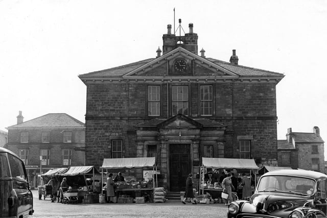Wetherby Town Hall on Market Place in April 1962. A virtually identical view can be seen on the Leodis website, but taken about 60 years earlier. In contrast, the siren has replaced the bell for summoning the Fire Brigade in this image. Motor cars have replaced the carts and stonework of the building has darkened. The old tradition of Market Day has continued, however, and stalls look very similar to the way they did circa 1900.