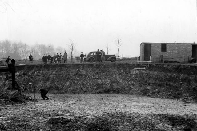Excavations in Cross Flatts Park in November 1942. A workman stands in the large hole, where the front of a digger is visible. On the rim of the hole are members of the public and a parked car.